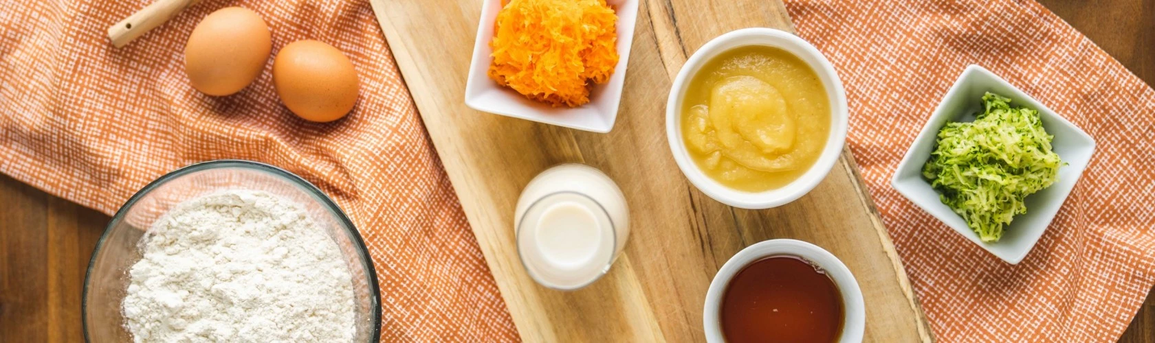 Draufsicht auf Backzutaten in Schüsseln auf Holzbrett und orangefarbenem Tuch, bereit zum Mischen.