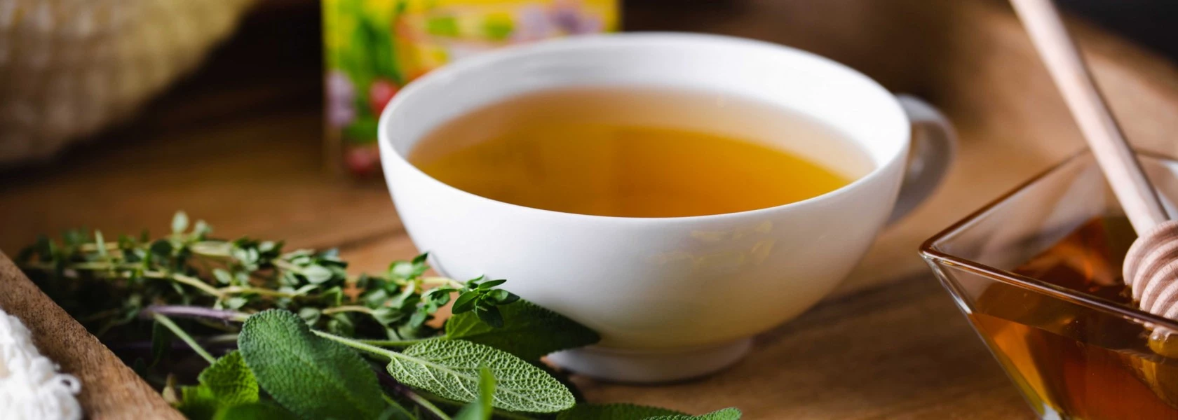Weißer Kräutertee mit frischen Kräutern, Honigschale und Schöpflöffel auf einer Holzoberfläche.