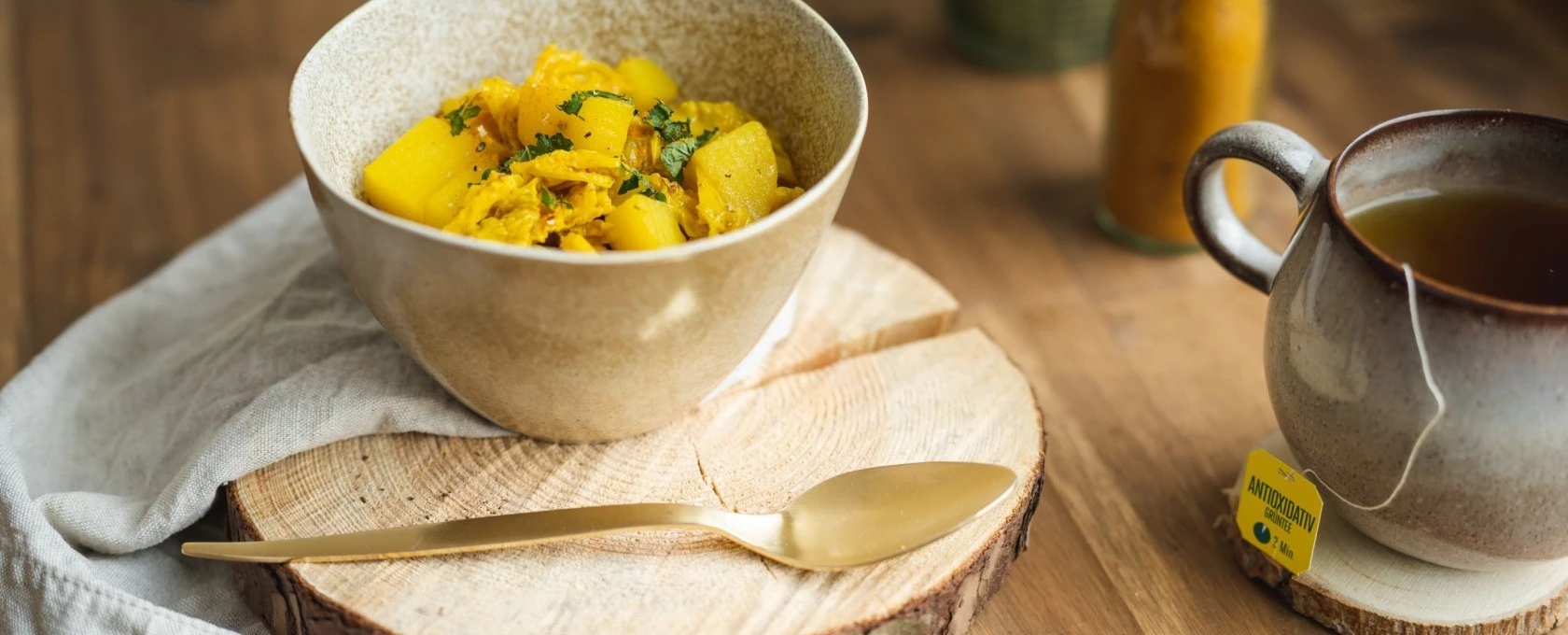 Beige Schüssel Hühnercurry mit Tee, goldenem Löffel und Serviette auf einem Holztisch.