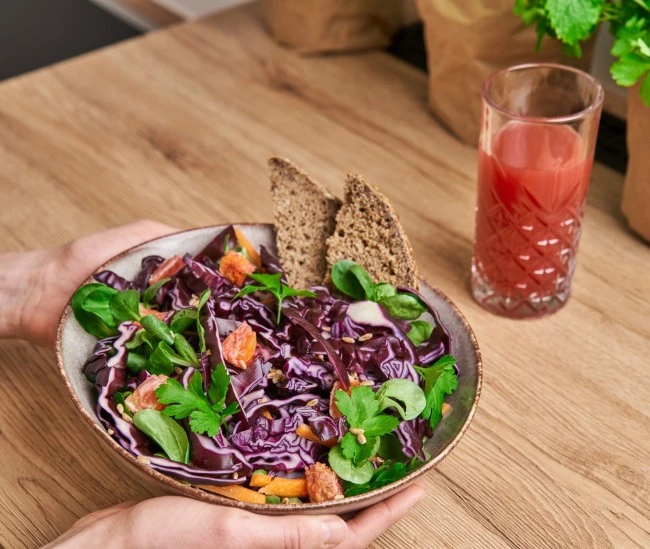 Person hält eine Salatschüssel mit Rotkohl, Gemüse und Brot; auf dem Tisch steht ein Glas rosa Saft.