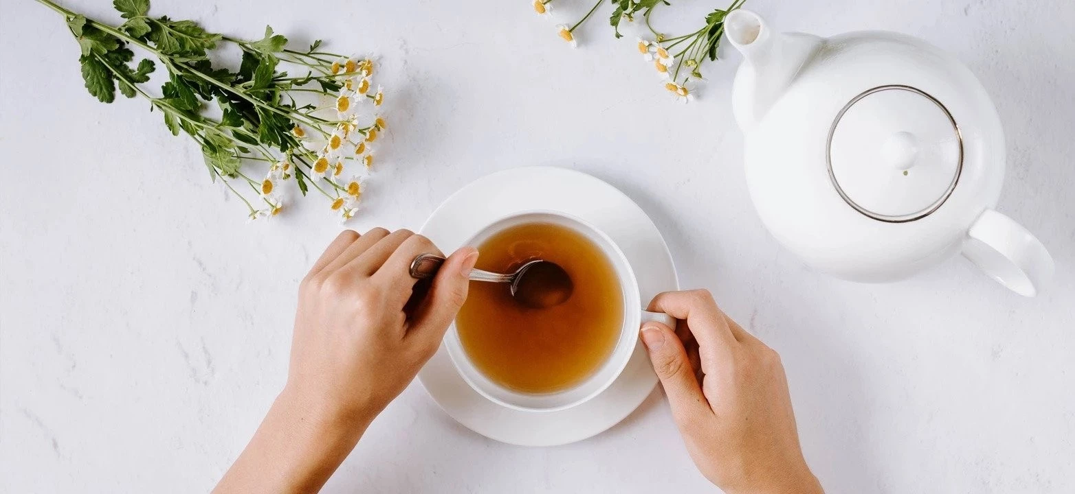 Person rührt Tee in Tasse um; weiße Teekanne und Kamillenblüten stehen auf weißer Fläche in der Nähe.