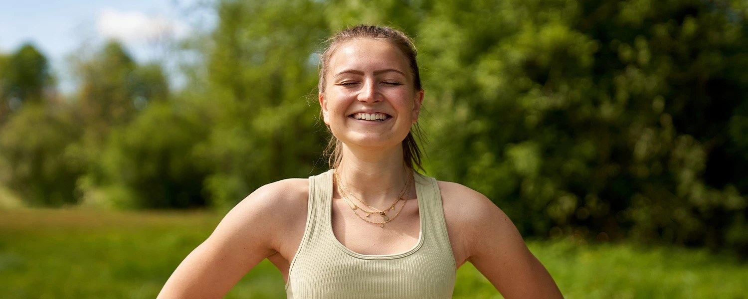 Lächelnde junge Frau im Freien, Augen geschlossen, trägt beiges Tanktop und mehrreihige Halsketten; Naturhintergrund.