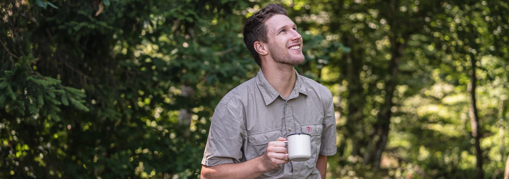 Lächelnder Mann im Freien, weiße Tasse in der Hand, umgeben von grünen Bäumen, durch deren Blätter das Sonnenlicht fällt.