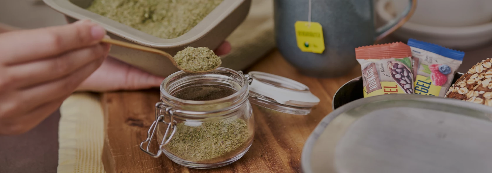Hand schöpft grünes Pulver in ein Glas neben einer Tasse mit Teebeutel und verschiedenen Snackriegeln.