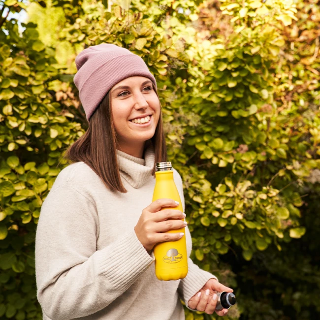 Lächelnde Frau mit rosa Mütze hält im Freien eine gelbe Wasserflasche, im Hintergrund grüne Büsche.