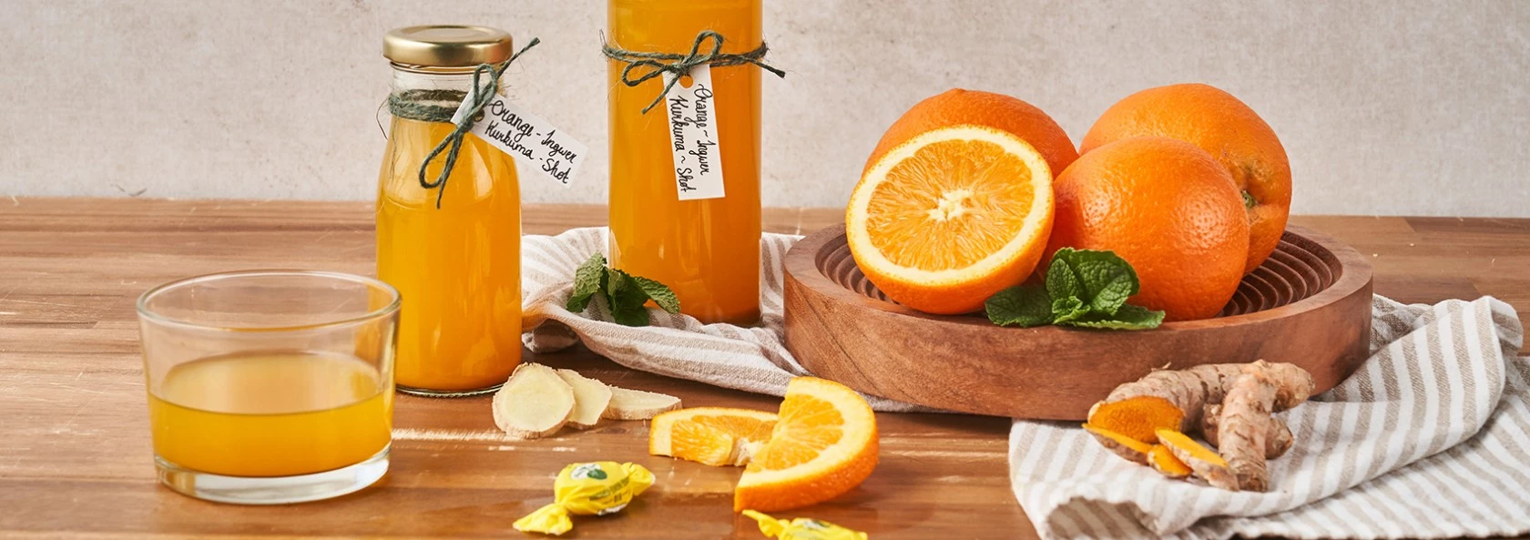 Orangensaft mit frischen Orangen, Ingwer, Kurkuma und Minze auf einem Holztisch neben gestreiftem Tuch.