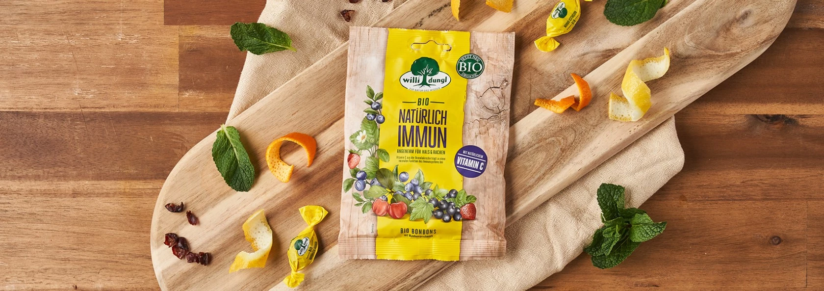 Wild Land Bio-Bonbonpackung auf Holzbrett mit unverpackten Bonbons, Minzblättern, Orangenschalen, Beeren.