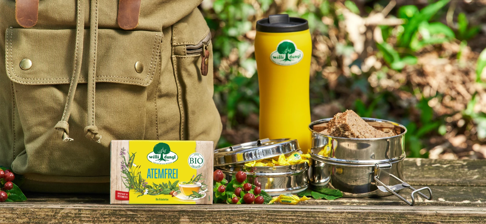 Picknickutensilien – gelbe Thermoskanne, Lunchboxen, Teebeutel, grüner Rucksack – auf einer Bank inmitten von Grün und Beeren.