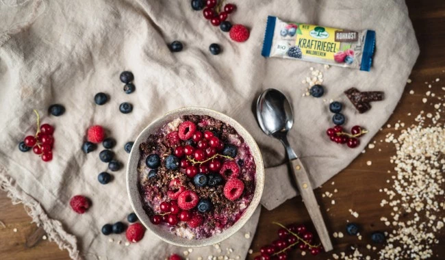 Smoothie-Schüssel mit Beeren, Löffel und Snackriegel auf beigem Tuch, Beeren verstreut.
