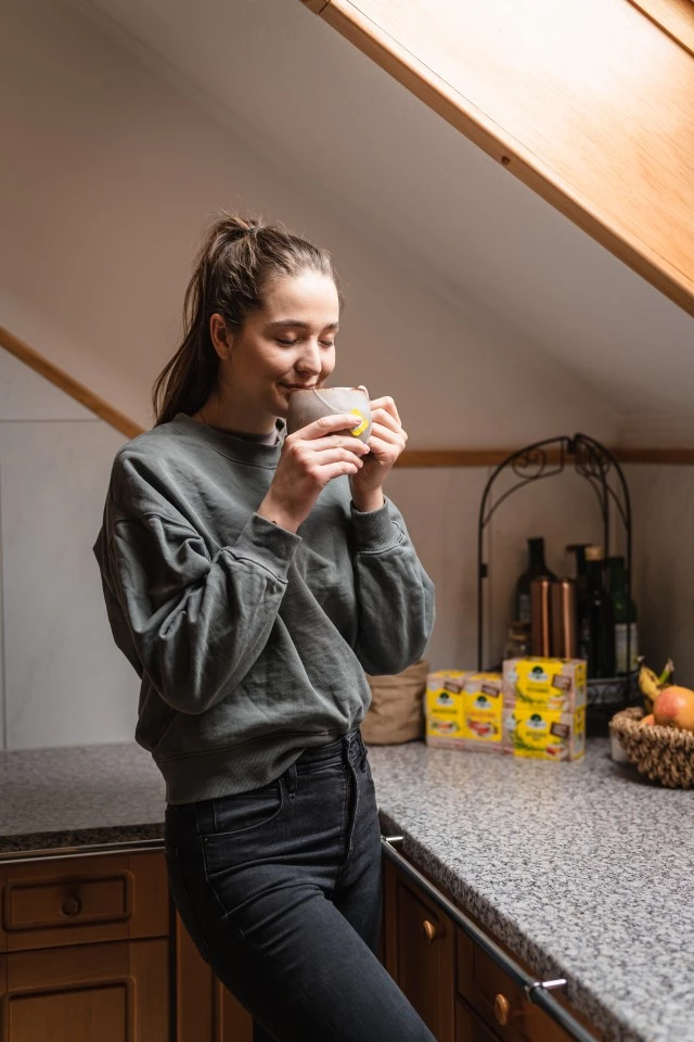 Lächelnde Frau genießt Tee in der Küche, Lipton-Packungen auf der Theke, Sonnenlicht strömt durch das Fenster.
