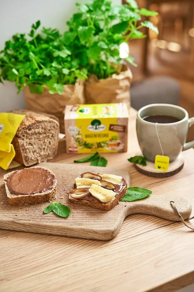 Vollkornbrot mit Schokoladenaufstrich und Banane, Tee, Minzblättern, Laib und Topfkräutern.