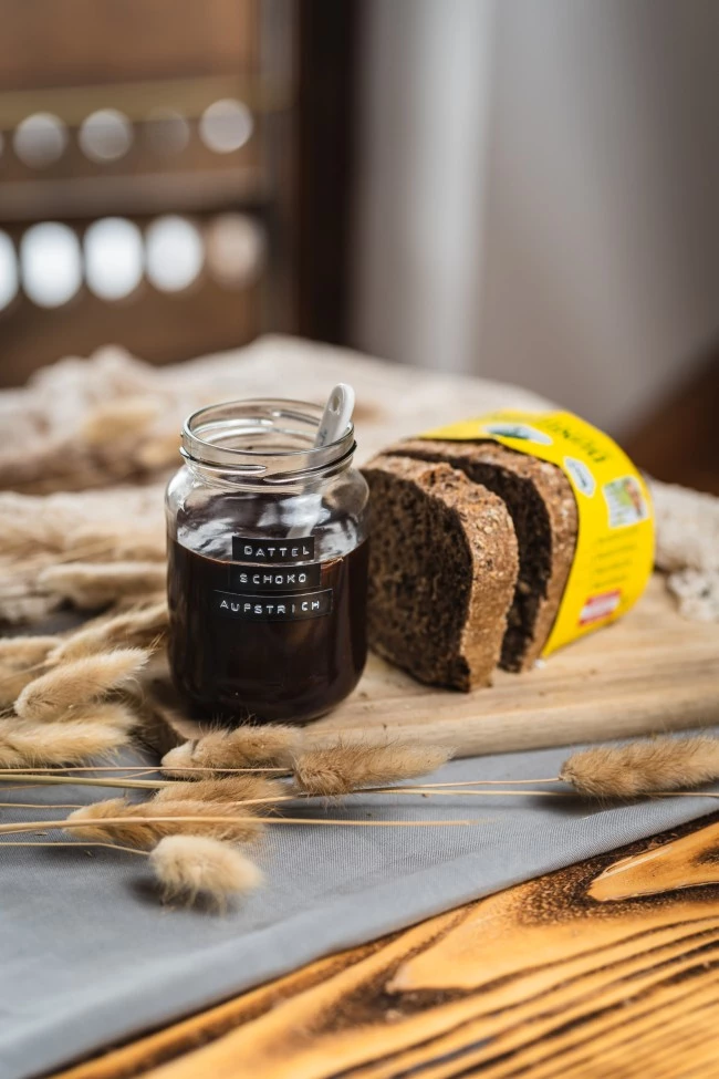 Glas Schokoladenaufstrich mit Löffel, geschnittenes Schwarzbrot, Weizenhalme auf Holzbrett.