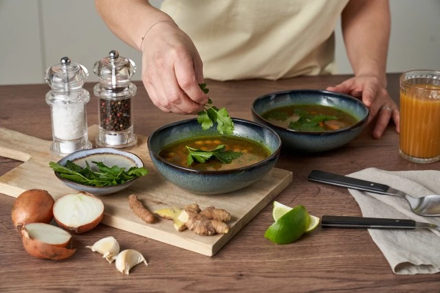 Person garniert Suppe mit frischen Kräutern; Zutaten, Utensilien und Saft sind auf einem Holztisch angeordnet.