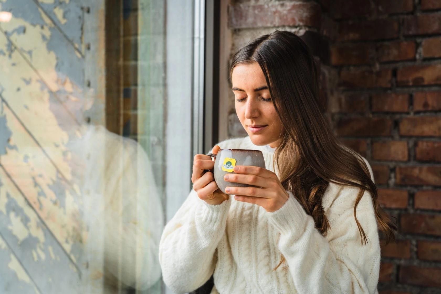 Frau im weißen Pullover hält eine Tasse am Fenster, die Augen geschlossen, wirkt ruhig; im Hintergrund eine Ziegelwand.