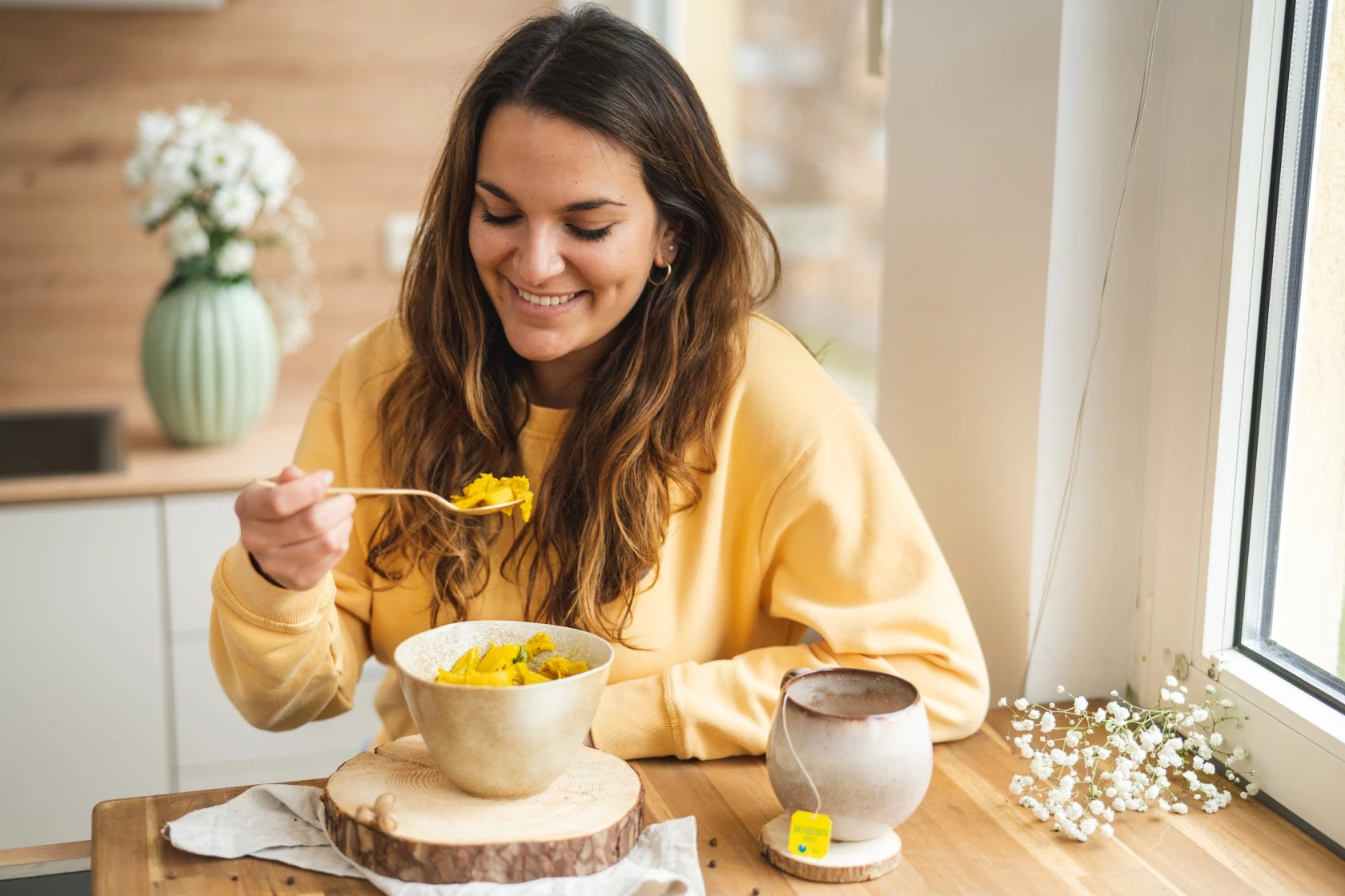 Frau im gelben Pullover isst lächelnd am Fenster, mit einer Teetasse und weißen Blumen auf dem Tisch.