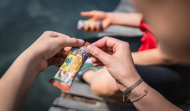 Hände packen den Kraftriegel-Snack am Steg aus, eine weitere Person isst in der Nähe am Wasser.