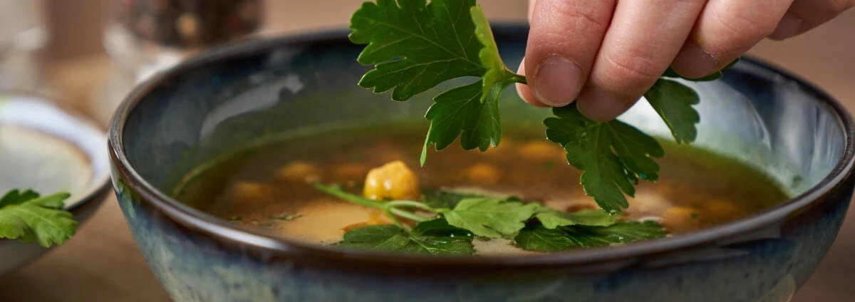 Hand garniert Suppe mit Petersilie in blau-grüner Keramikschale, Kichererbsen und Kräuter auf Holztisch sichtbar.