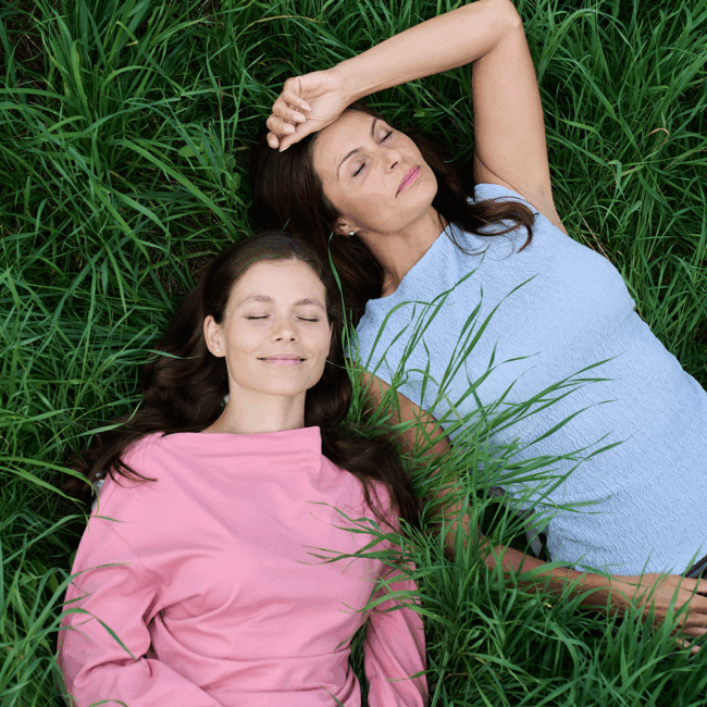 Zwei Frauen liegen nebeneinander im hohen Gras, die Augen geschlossen, eine lächelt sanft.