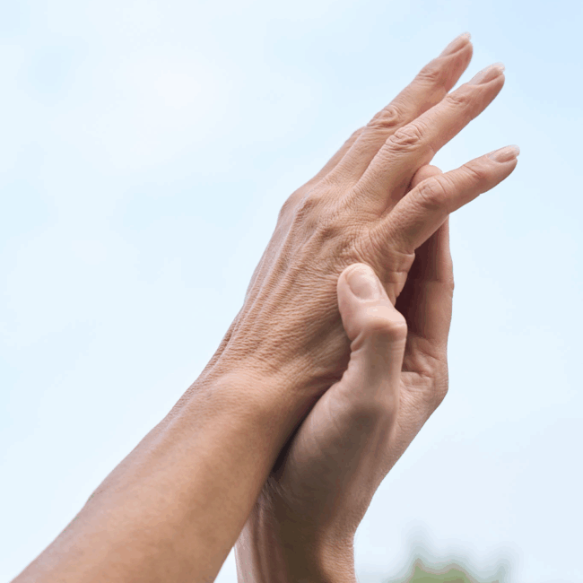 Zwei Hände berühren sich sanft vor einem hellblauen Himmel, Adern und natürliche Hautstruktur sind sichtbar.