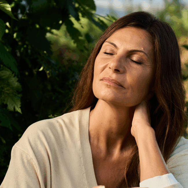Frau mit langen braunen Haaren sitzt im Freien im Sonnenlicht, Augen geschlossen, friedlich und entspannt wirkend.