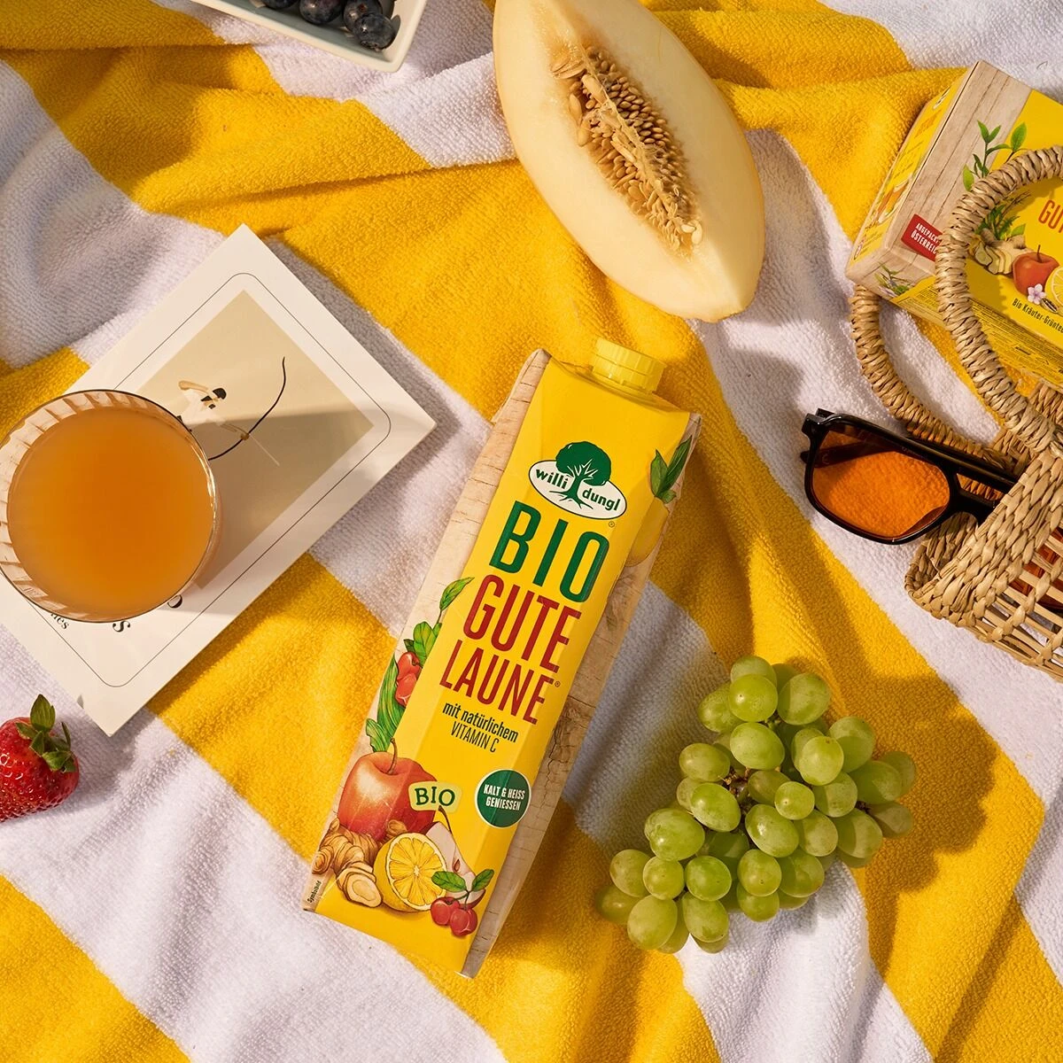Ein Karton „Gute Laune Bio“-Saft, ein Glas Orangensaft, Weintrauben, eine Melonenscheibe, eine Erdbeere und eine Sonnenbrille, angeordnet auf einem gelb-weiß gestreiften Handtuch.