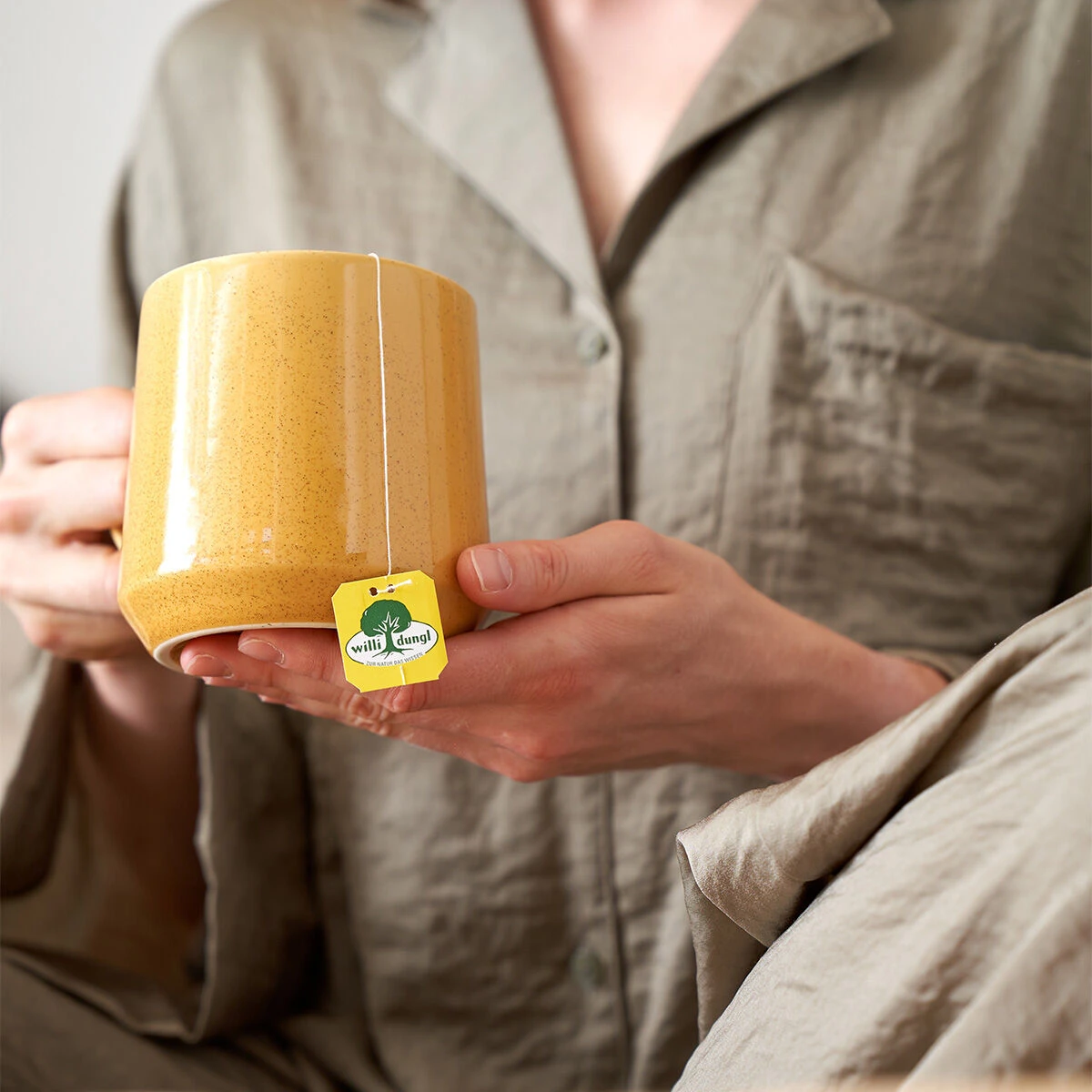 Eine Person im hellbraunen Pyjama hält eine große gelbe Tasse Tee mit einem sichtbaren gelben Etikett mit der Aufschrift „Wach & Konzentriert“ auf der Teebeutelschnur.