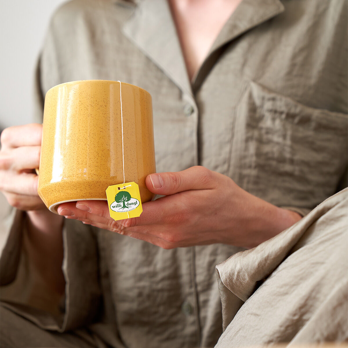 Eine Person im hellgrauen Pyjama hält eine große gelbe Tasse mit einem Teebeutel mit der Aufschrift „9 Kräuter BIO“ in der Hand und sorgt so für eine entspannte und gemütliche Atmosphäre.