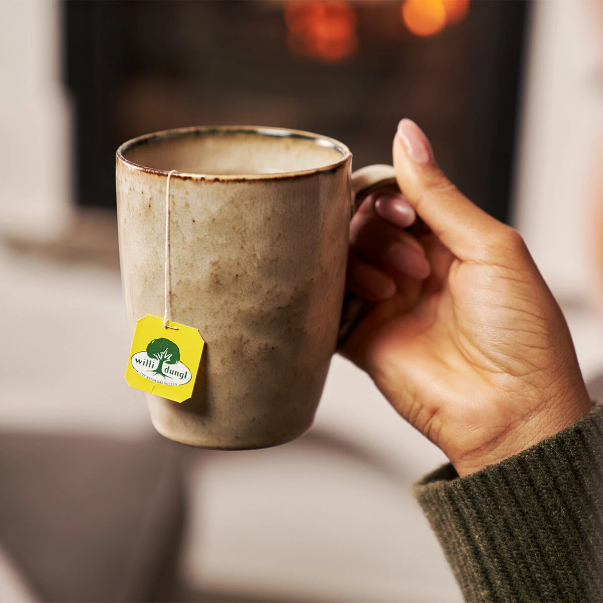 Eine Hand in einem grünen Pullover hält eine beige Keramiktasse mit einem Bergkräuter BIO-Teebeutel vor einem verschwommenen, warmen Kaminfeuer-Hintergrund.