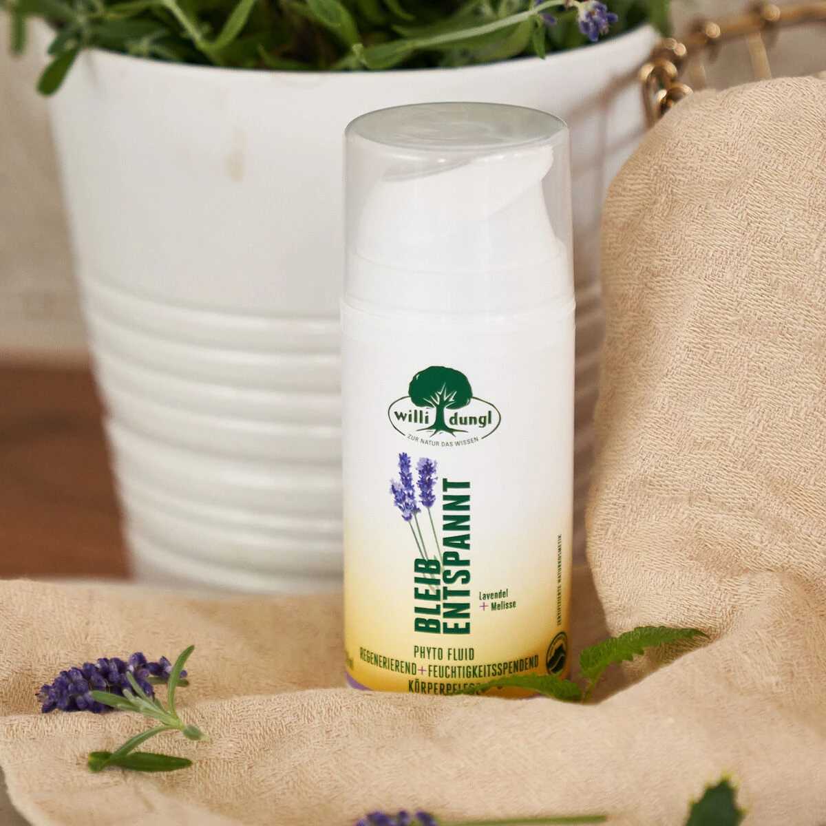 Eine Flasche BLEIB ENTSPANNT Phyto-Fluid 100ml liegt auf einem beigen Handtuch, daneben befinden sich Lavendelzweige, während im Hintergrund ein weißer Blumentopf und üppige grüne Blätter zu sehen sind.