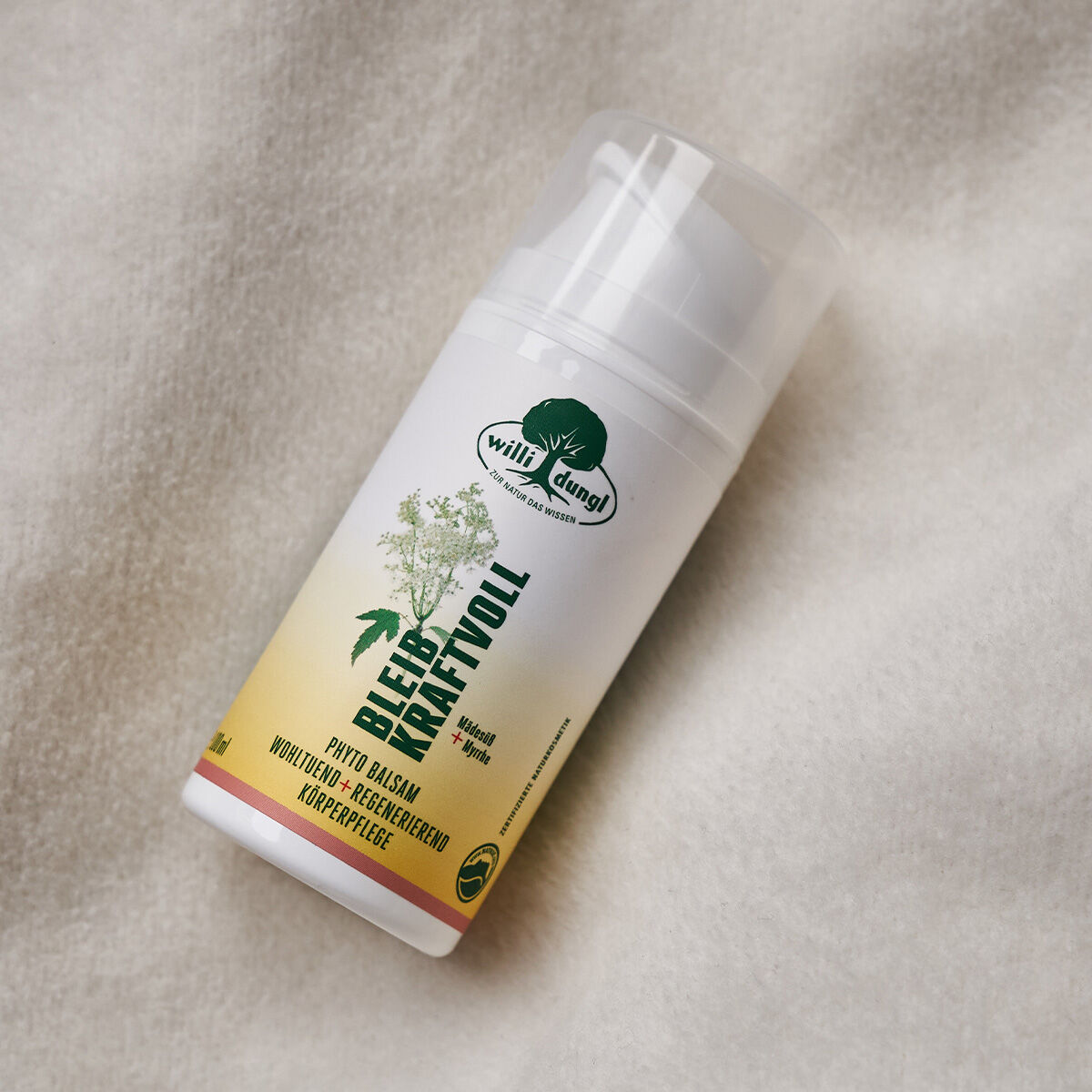 Eine 100-ml-Flasche BLEIB KRAFTVOLL Phyto-Balsam mit einem weiß-gelben Etikett, grünem Text und einer Pflanzenillustration ruht auf einem weichen, weiß strukturierten Stoff.