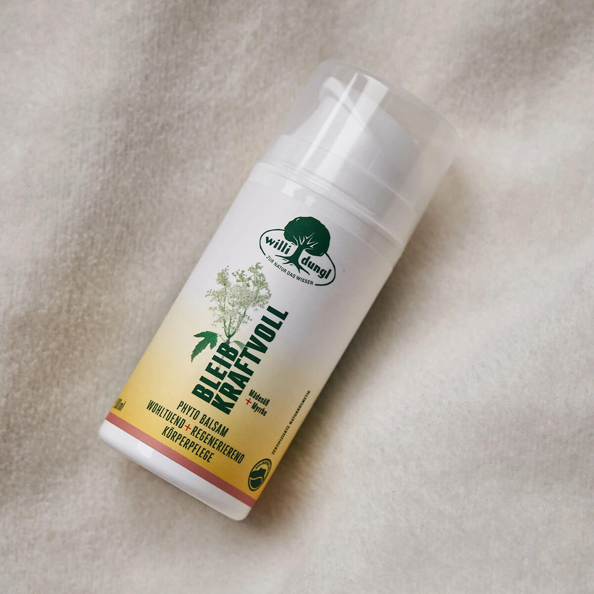 Eine 100-ml-Flasche BLEIB KRAFTVOLL Phyto-Balsam mit einem weiß-gelben Etikett, grünem Text und einer Pflanzenillustration ruht auf einem weichen, weiß strukturierten Stoff.