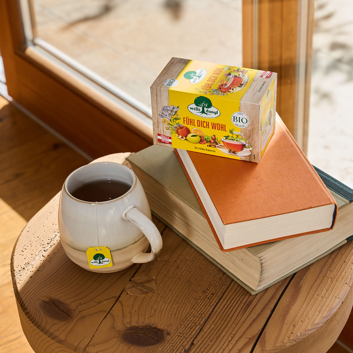 Auf einem Holzhocker steht neben zwei Büchern eine Tasse Tee, darauf eine Schachtel „Fühl dich wohl BIO“-Kräutertee. Sonnenlicht durch das Fenster sorgt für eine gemütliche Atmosphäre.