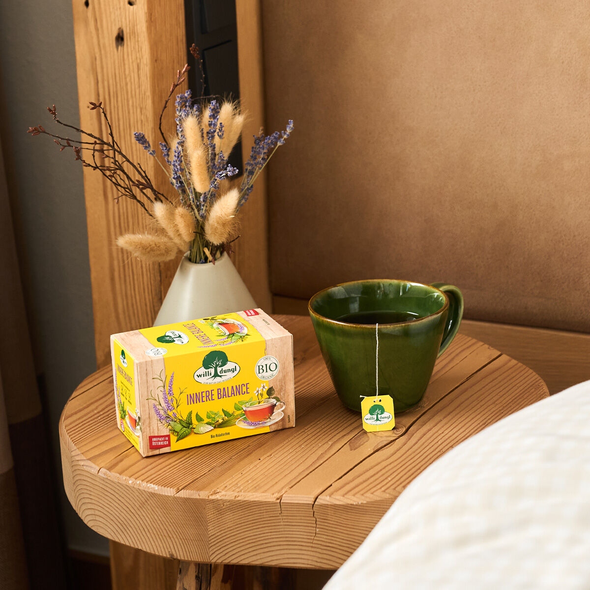 Eine grüne Tasse Tee und eine Schachtel Innere Balance BIO-Kräutertee stehen auf einem Nachttisch aus Holz neben einer kleinen Vase mit getrockneten Blumen und einem beigefarbenen Polsterbett.
