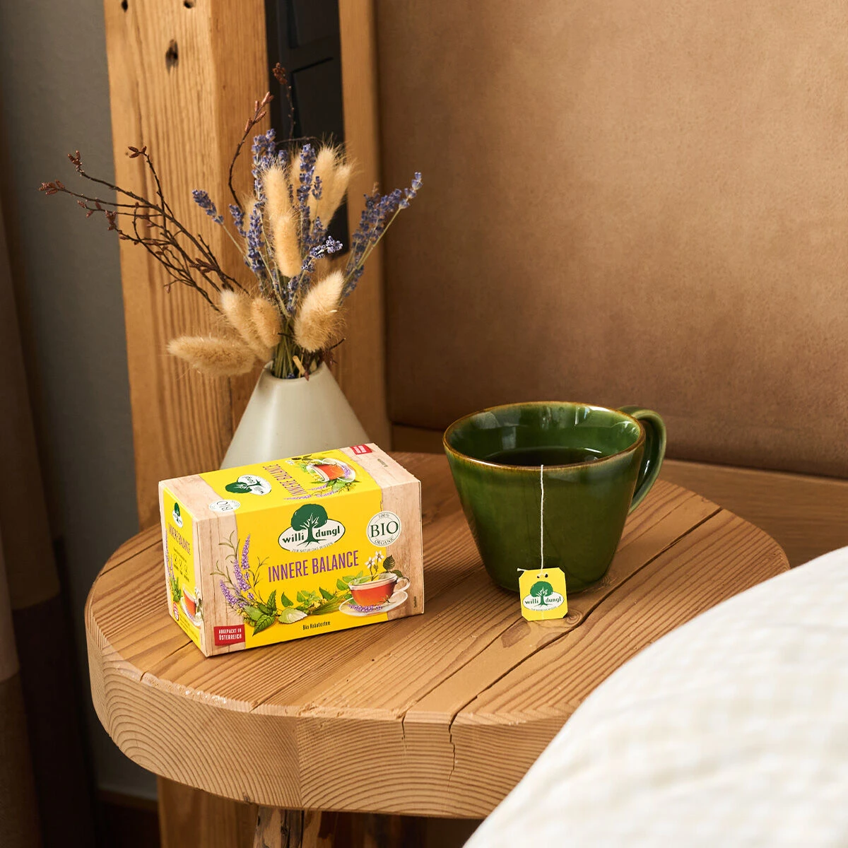 Eine grüne Tasse Tee und eine Schachtel Innere Balance BIO-Kräutertee stehen auf einem Nachttisch aus Holz neben einer kleinen Vase mit getrockneten Blumen und einem beigefarbenen Polsterbett.