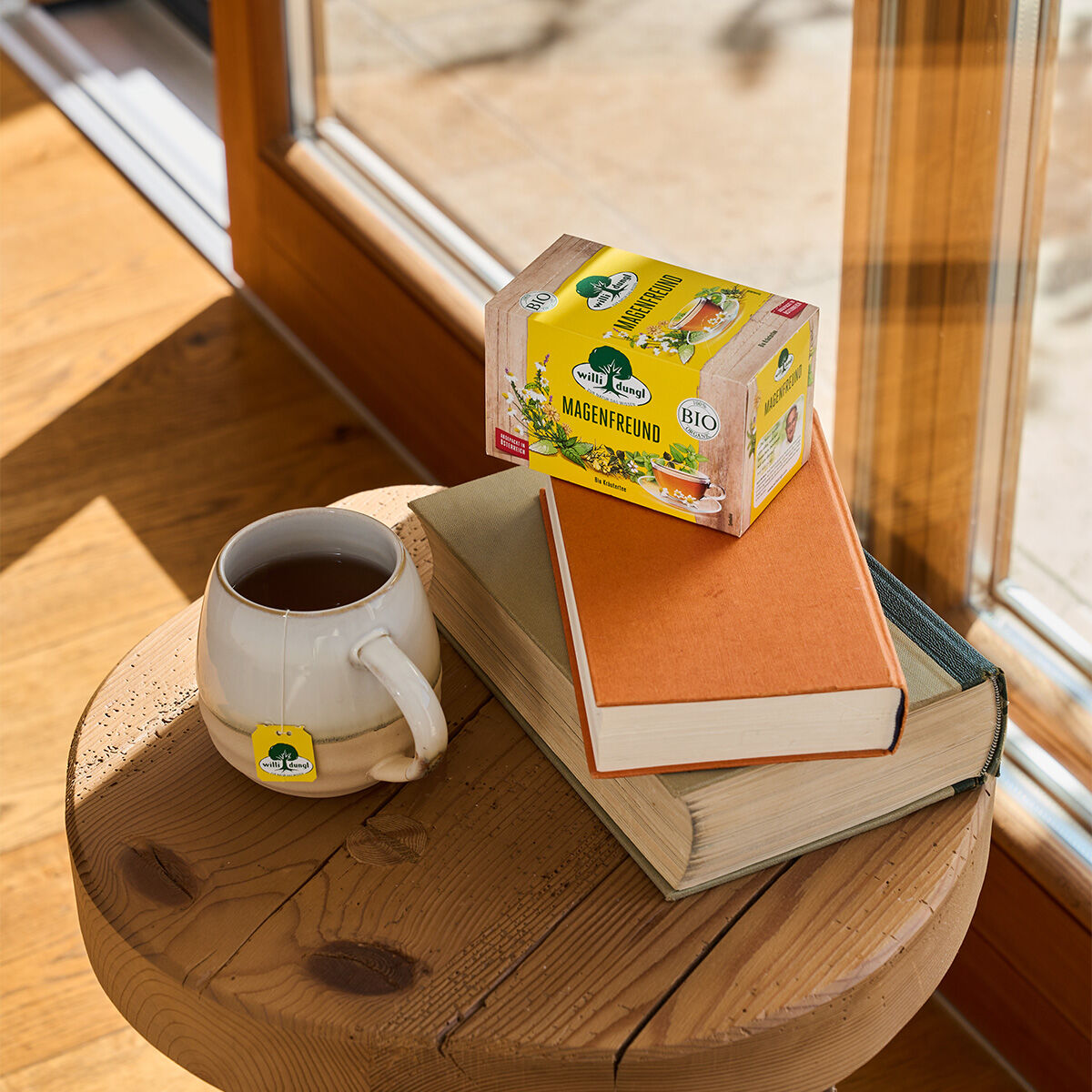 Auf einem Holzhocker an einem sonnigen Fenster stehen zwei Bücher, eine Tasse Magenfreund BIO-Tee mit dem Etikett am Henkel und eine Schachtel Magenfreund BIO, während warmes Sonnenlicht durch das Glas strömt.