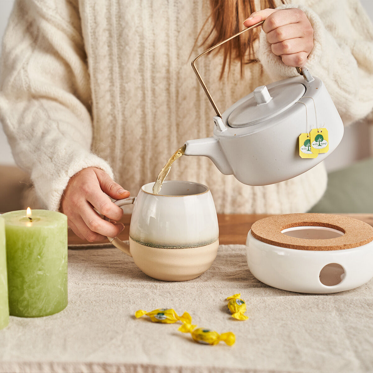 Eine Person im cremefarbenen Pullover gießt Tee aus einer weißen Teekanne in eine Tasse. Daneben stehen eine grüne Kerze, eingewickelte Bonbons und ein Stövchen – perfekt für gemütliche Momente mit Natürlich Immun BIO.