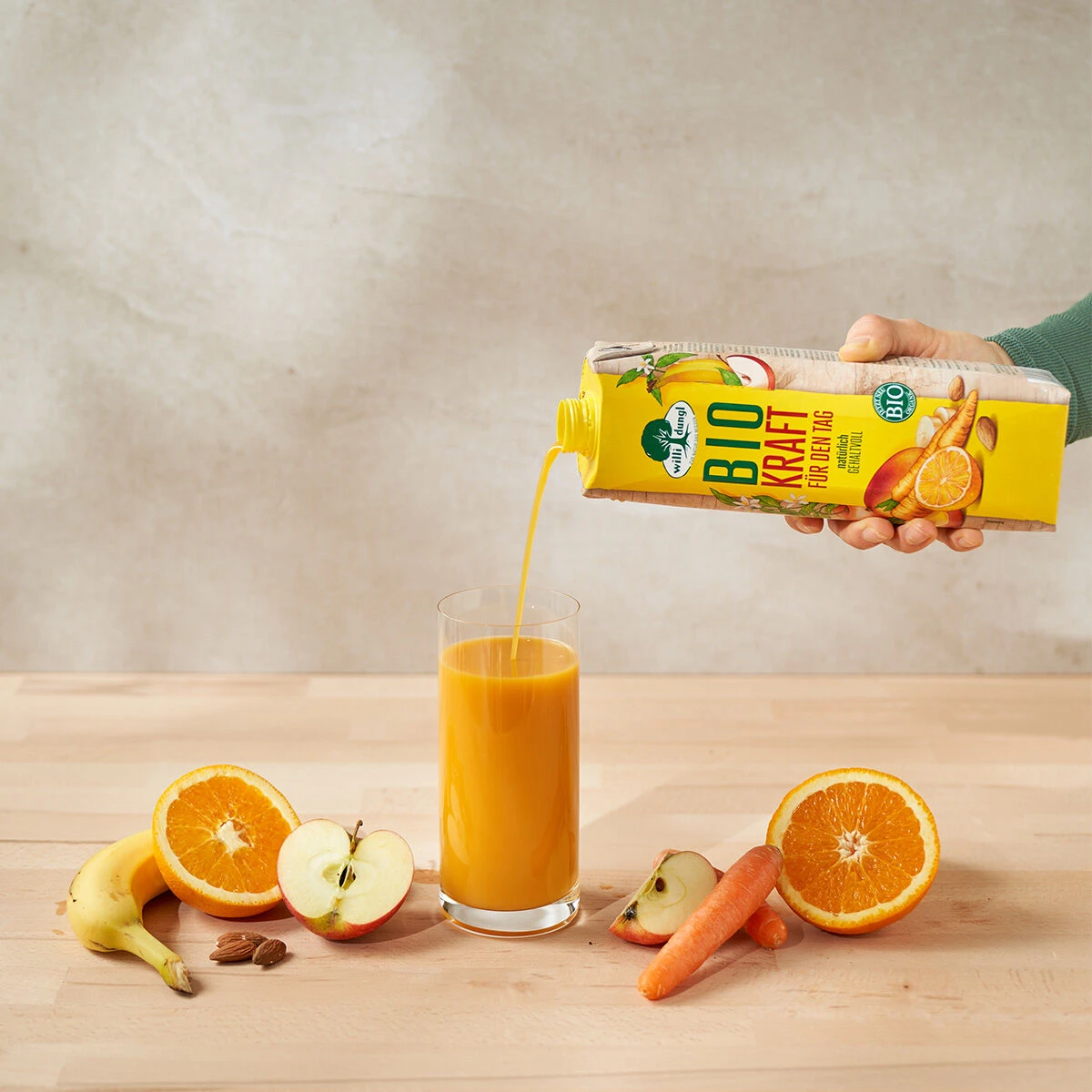 Eine Hand gießt Orangensaft aus einem gelben Kraft für den Tag Bio-Karton in ein Glas. Frische Banane, Apfel, Karotte und Orange werden für Kraft für den Tag auf einer hellen Holzfläche arrangiert.