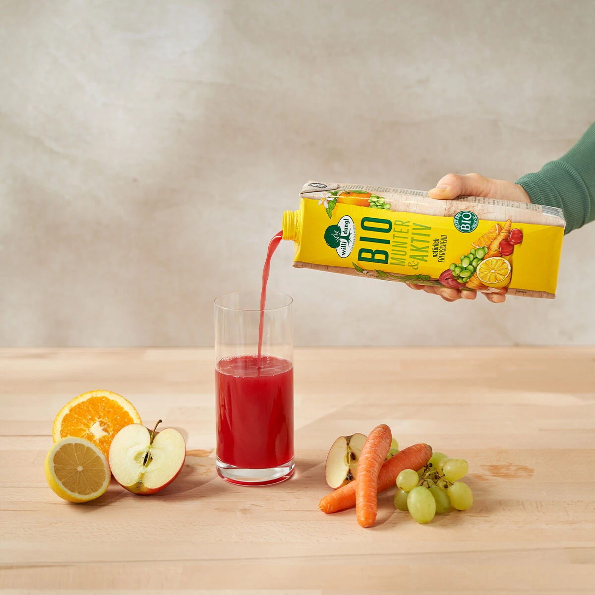 Eine Hand gießt Munter & Aktiv Bio aus einem gelben Karton in ein Glas. Auf dem Tisch stehen eine Orangenhälfte, Apfelscheiben, Zitrone, Karotten und grüne Trauben. Der Hintergrund ist neutral und hell.