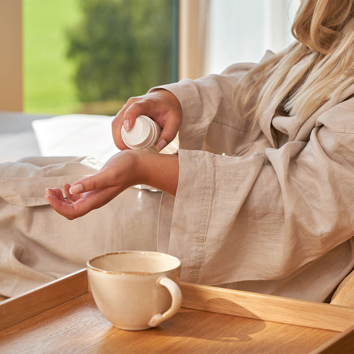 In einem beigen Bademantel trägt jemand am Fenster die VERWÖHNE DICH Phyto-Lotion 200ml auf sein Handgelenk auf. Eine Keramiktasse auf einem Holztablett unterstreicht die ruhige, entspannte Morgenstimmung.