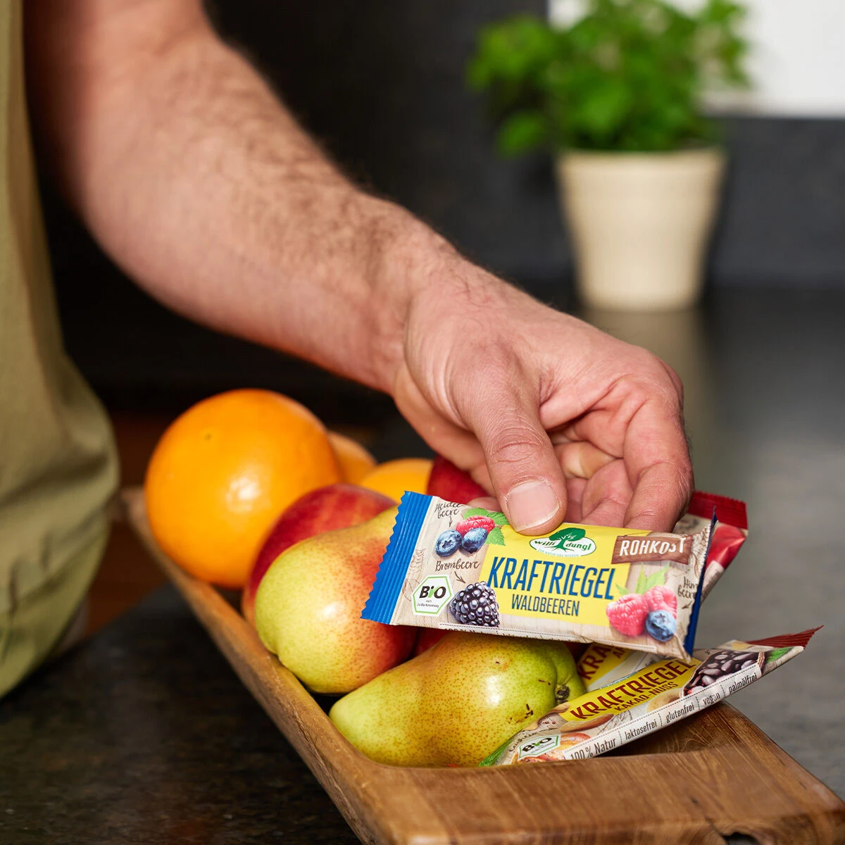 Eine Hand greift in eine hölzerne Obstschale gefüllt mit Birnen, Orangen und Nektarinen und nimmt einen Kraftriegel Waldbeeren BIO Energieriegel heraus.